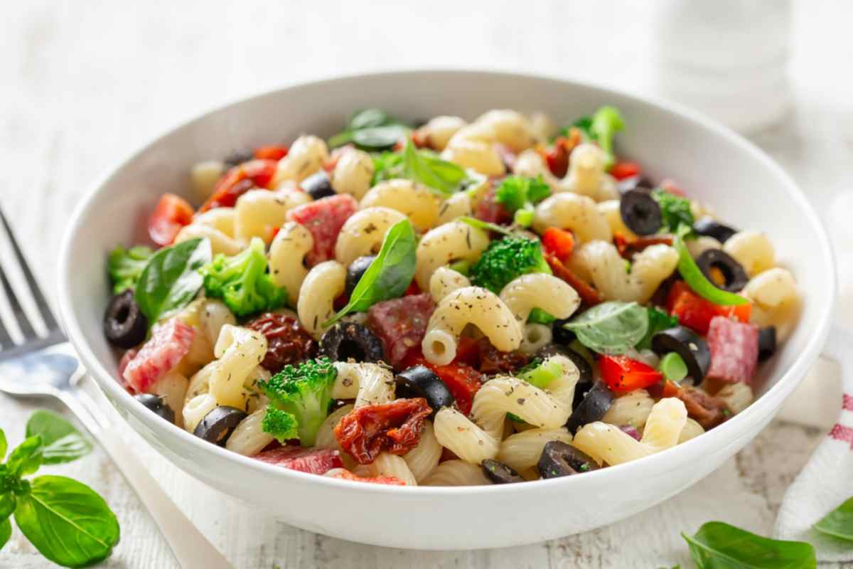 Colorful pasta salad with olives, broccoli, peppers, and herbs in a white bowl.