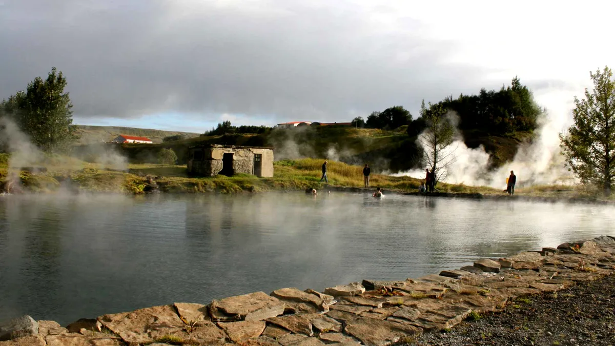 Bañistas en la Secret Lagoon de Islandia con vapor y caseta de piedra al fondo.