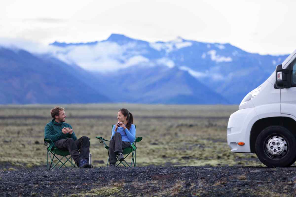 Camping en Islandia Pareja descansando en sillas plegables junto a una autocaravana; llanura volcánica y montañas nevadas al fondo, pausa de café durante el viaje.