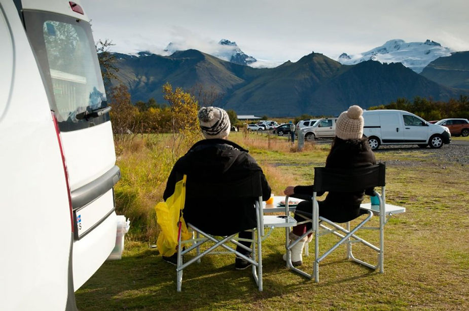 Coppia a un tavolino accanto al camper nel campeggio di Skaftafell, vista su montagne e ghiacciai del Vatnajökull.