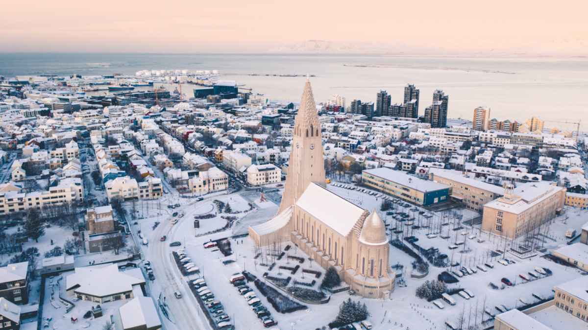 Guida Reykjavík Vista aerea di Reykjavík innevata con Hallgrímskirkja al centro, strade imbiancate e oceano sullo sfondo in una chiara mattina d’inverno.