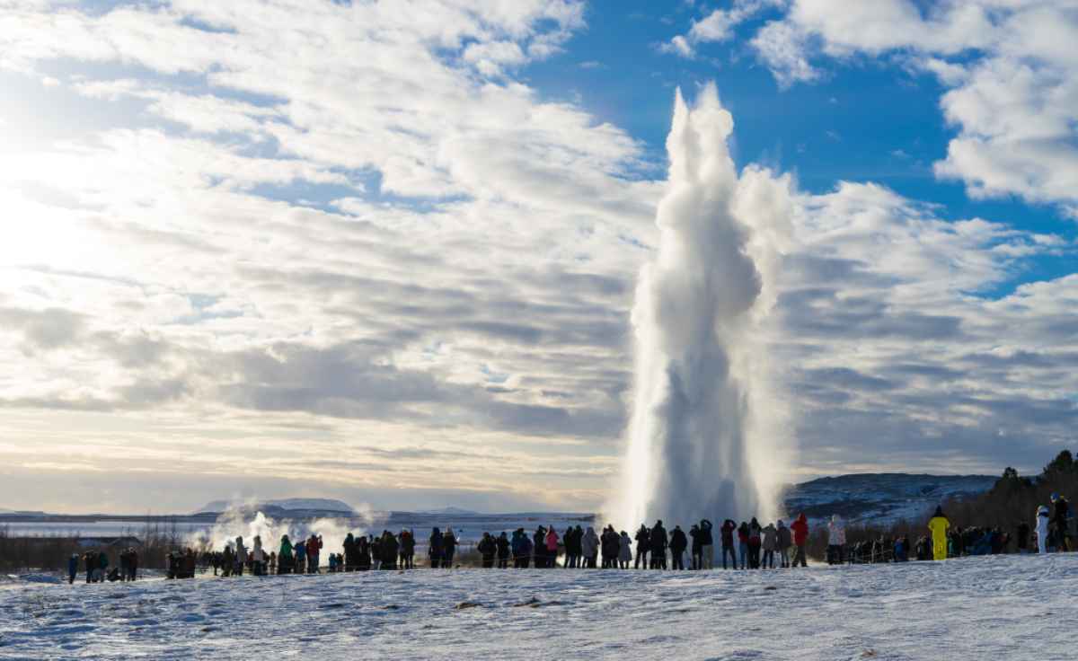 Strokkur in inverno Geyser Strokkur erutta tra neve e nuvole; gruppo di visitatori osserva da lontano in una fredda giornata invernale nel Golden Circle.