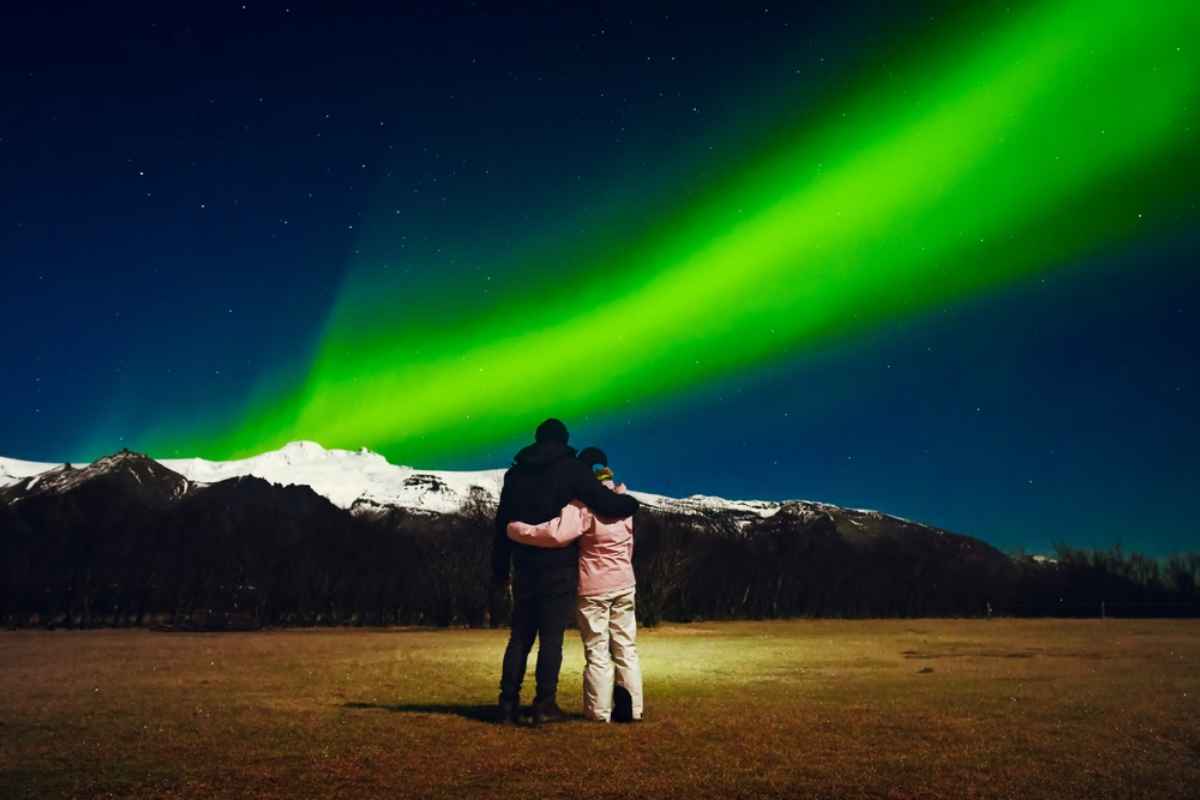 Due persone abbracciate osservano un nastro verde di aurora boreale sopra montagne innevate, cielo stellato d’inverno in Islanda.