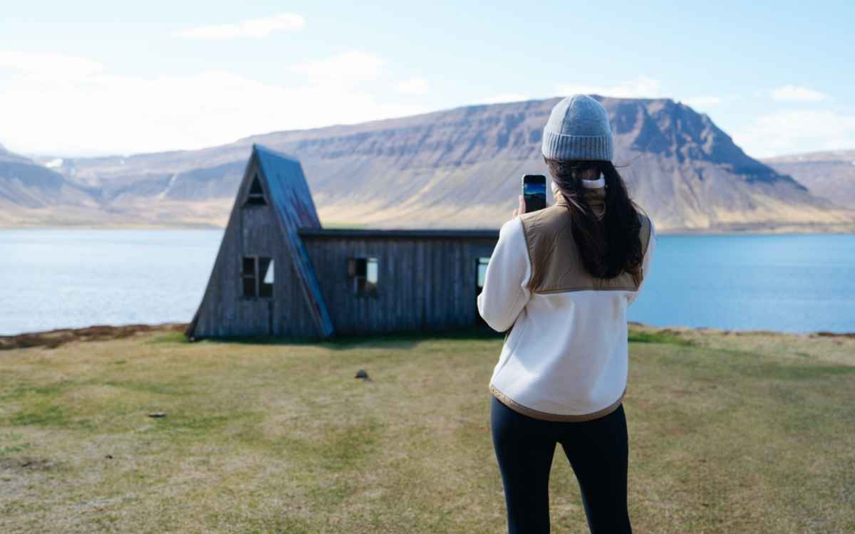 Viaggiatrice che fotografa una capanna a tetto a punta sulle rive di un fiordo islandese con montagne sullo sfondo.