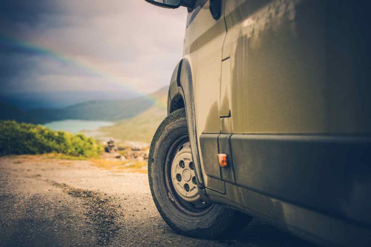 Ruota sotto arcobaleno Primo piano della ruota di un camper su strada di montagna con arcobaleno in lontananza.