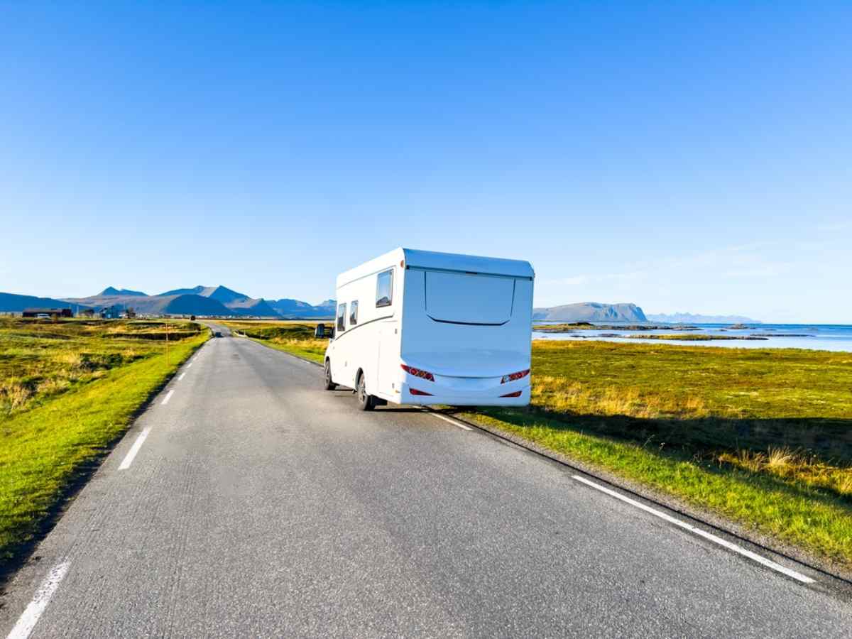 Camper sulla costa Autocaravan che percorre una strada costiera soleggiata con fiordi e montagne in lontananza.