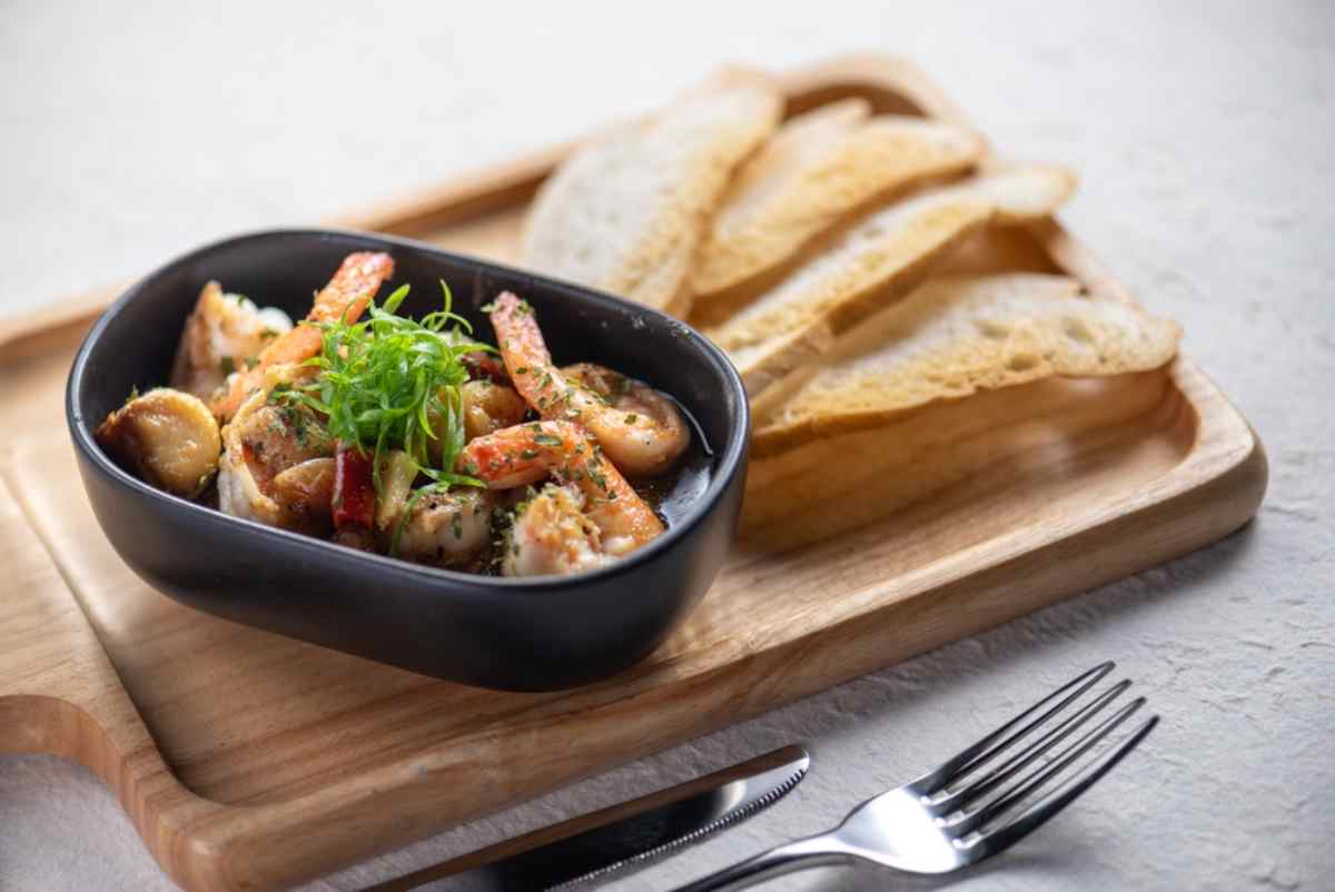 Pirofila con gamberi saltati all’aglio, erbe e fette di pane croccante su tagliere.