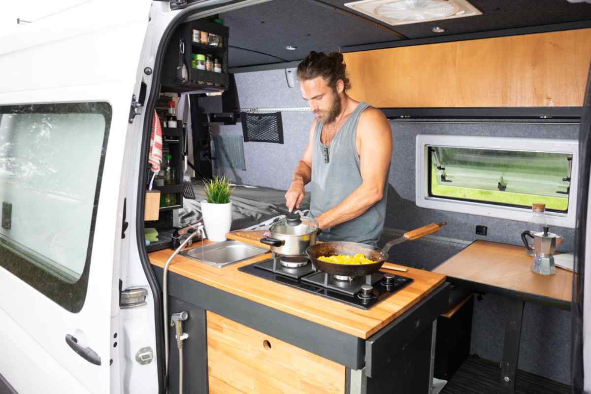 Viaggiatore che cucina nella cucina compatta del campervan, fornelli a gas e piano in legno.