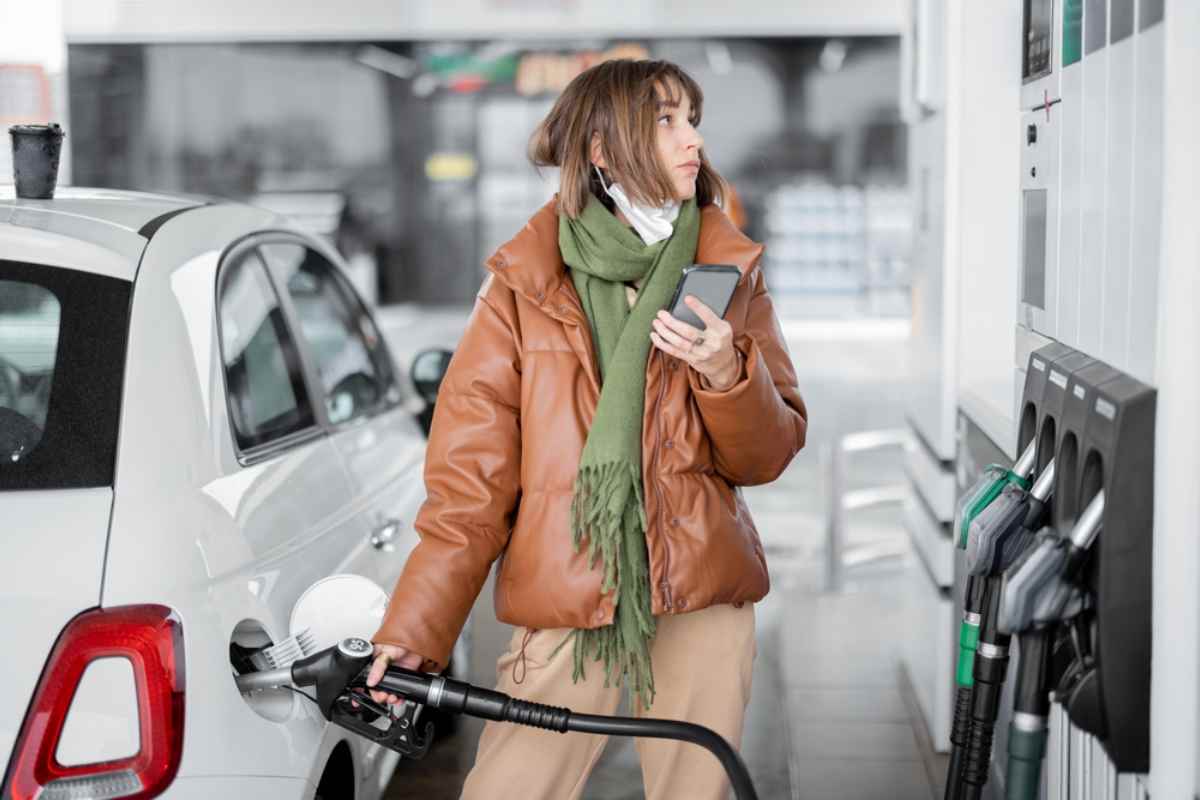 Calcolare il prezzo della benzina in Islanda Una ragazza con cappotto in pelle marrone tiene la pistola del carburante con la mano destra mentre la inserisce nel tappo del veicolo per fare rifornimento; con la sinistra tiene il cellulare per calcolare il prezzo della benzina in Islanda.