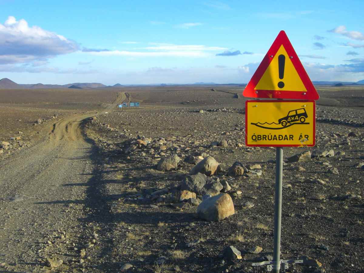 Yellow “Óbrúaðar ár” warning sign for unbridged rivers with a 4x4 icon beside a rough Highland track across a rocky, barren plain.