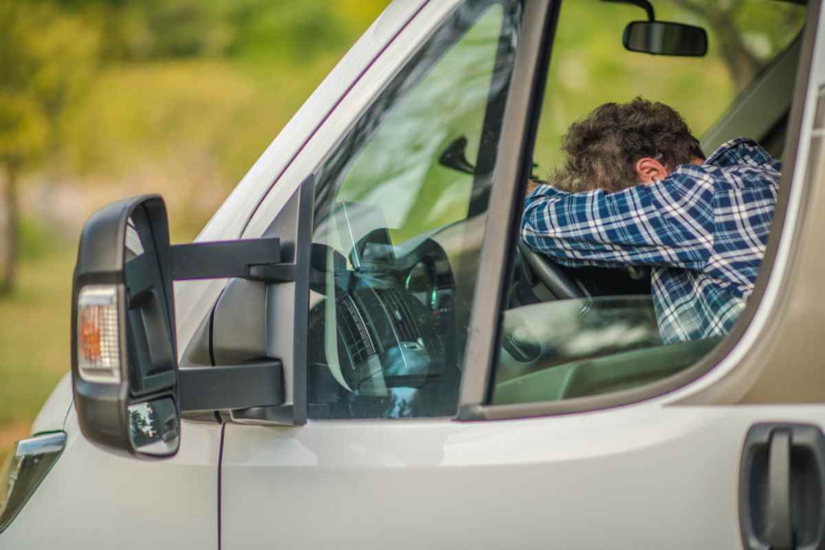 Tired driver in a campervan rests his head on the steering wheel, plaid shirt visible through the side window, green background softly blurred.