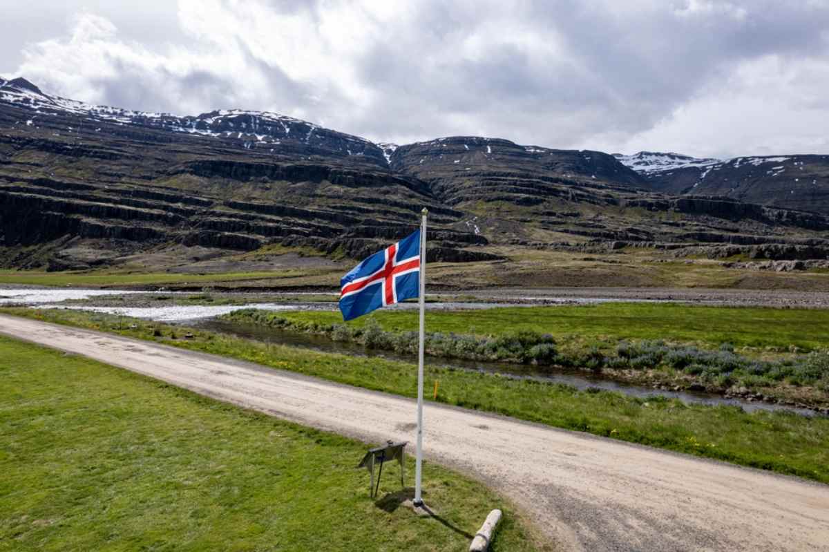 Icelandic flag waves beside a gravel track and small river in a wide mountain valley, layered cliffs and patches of snow under cloudy skies.