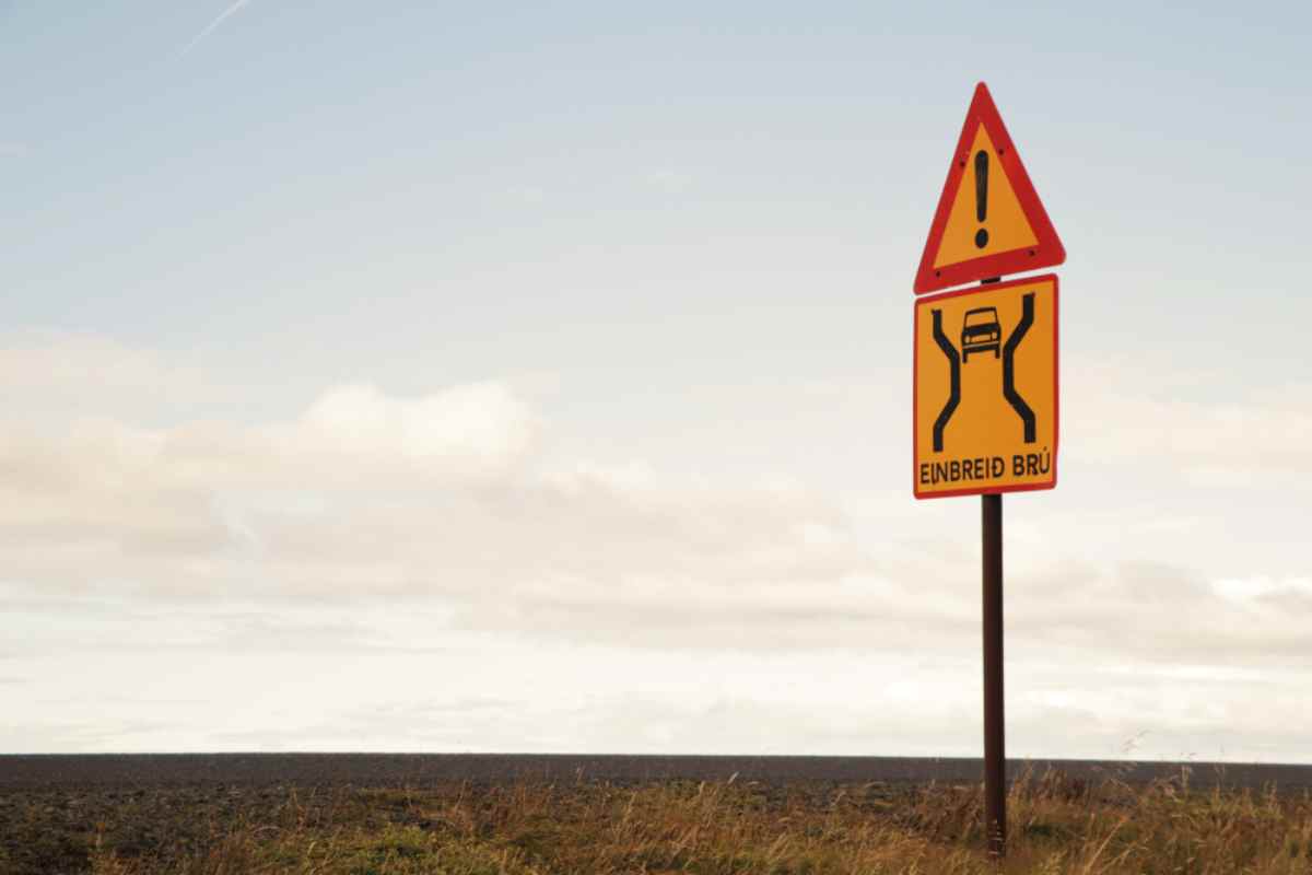 Yellow “Einbreið brú” single-lane bridge warning sign on a grassy roadside in Iceland, pale sky and open horizon behind.