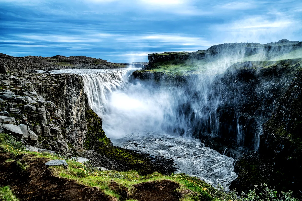 A breathtaking view of Dettifoss Waterfall in Iceland, known as Europe's most powerful waterfall. The massive cascade of water plunges into a rugged canyon, with mist rising from the turbulent waters below. The surrounding landscape is characterized by rocky cliffs and sparse vegetation under a vivid blue sky.