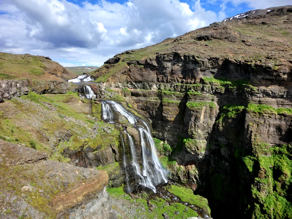 A stunning view of Glymur Waterfall in Iceland, one of the highest waterfalls in the country. The waterfall cascades down a series of rocky cliffs, surrounded by green moss and rugged terrain. The sky above is dotted with clouds, adding to the dramatic landscape. The distant snowy peaks are visible, creating a picturesque and awe-inspiring scene.