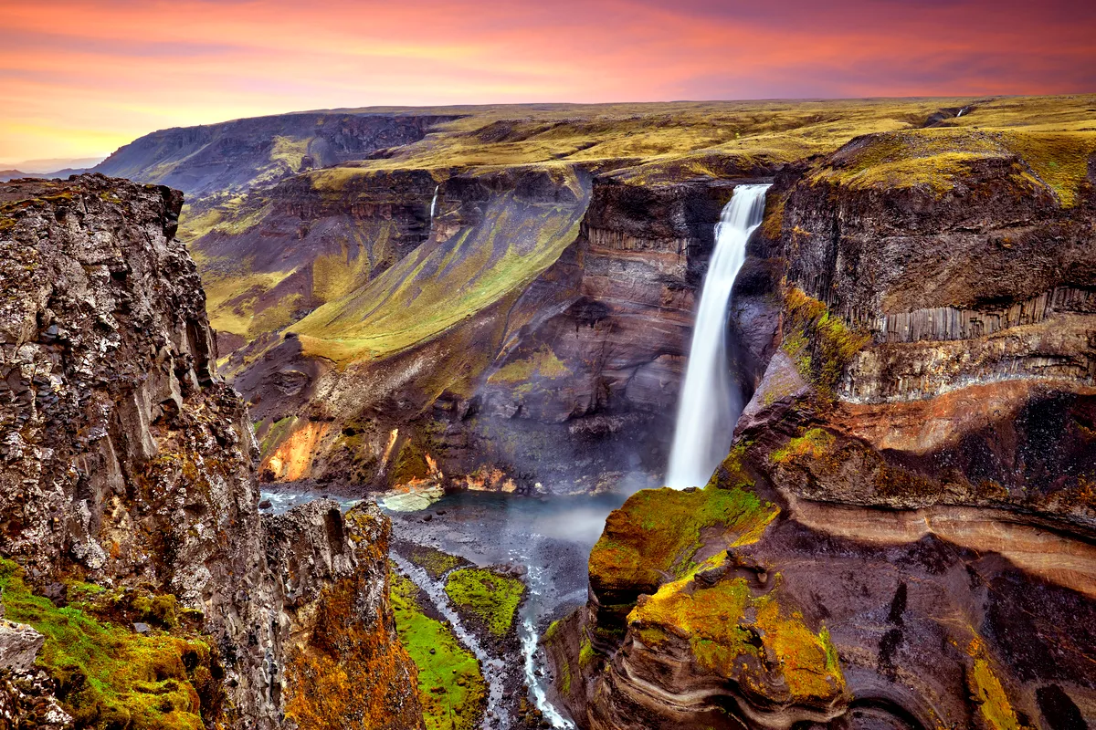 A stunning view of Haifoss Waterfall in Iceland at sunset. The waterfall plunges dramatically from a high cliff into a deep canyon, surrounded by rugged terrain with varying shades of green and brown. The sky above is painted in warm hues of orange and pink, enhancing the natural beauty of the landscape. The scene captures the raw power and serene majesty of one of Iceland's most breathtaking waterfalls.