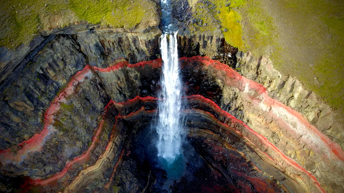 An aerial view of Hengifoss Waterfall in Iceland. The waterfall plunges down a sheer cliff face, surrounded by striking layers of red clay and basalt rock, creating a vivid contrast against the cascading white water. The rocky landscape around the waterfall is dotted with patches of green moss and vegetation. The waterfall descends into a deep pool at the base, creating a misty spray. This image captures the unique geological features and dramatic beauty of one of Iceland's tallest waterfalls.