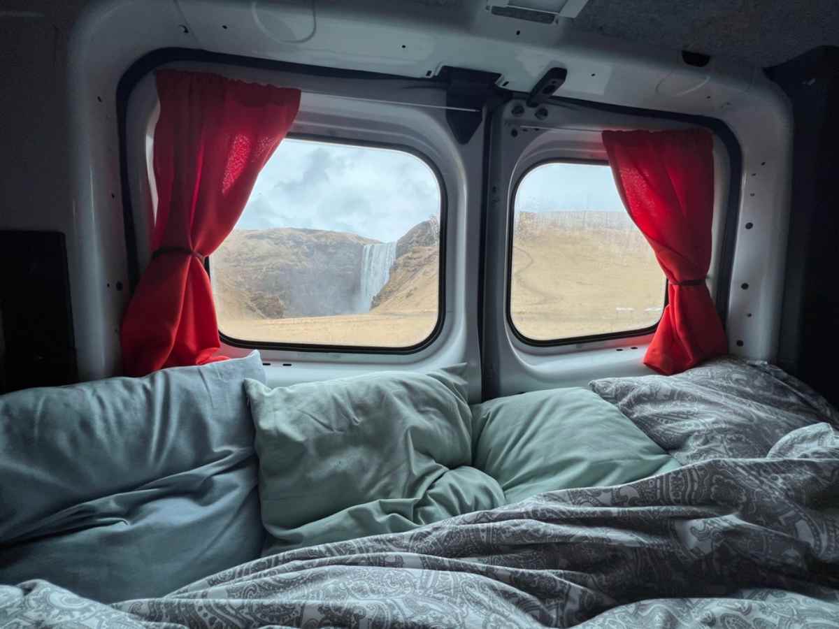 Image of the interior of the back of a campervan showing an unmade bed under windows showing an Icelandic waterfall in the distance.