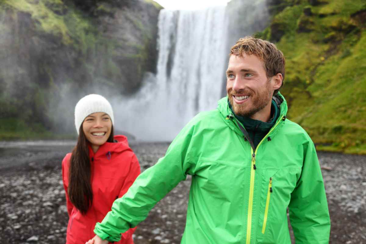 A hiking couple, man and wife, smilingly enjoy the experience of visiting a waterfall in Iceland, visible behind them.