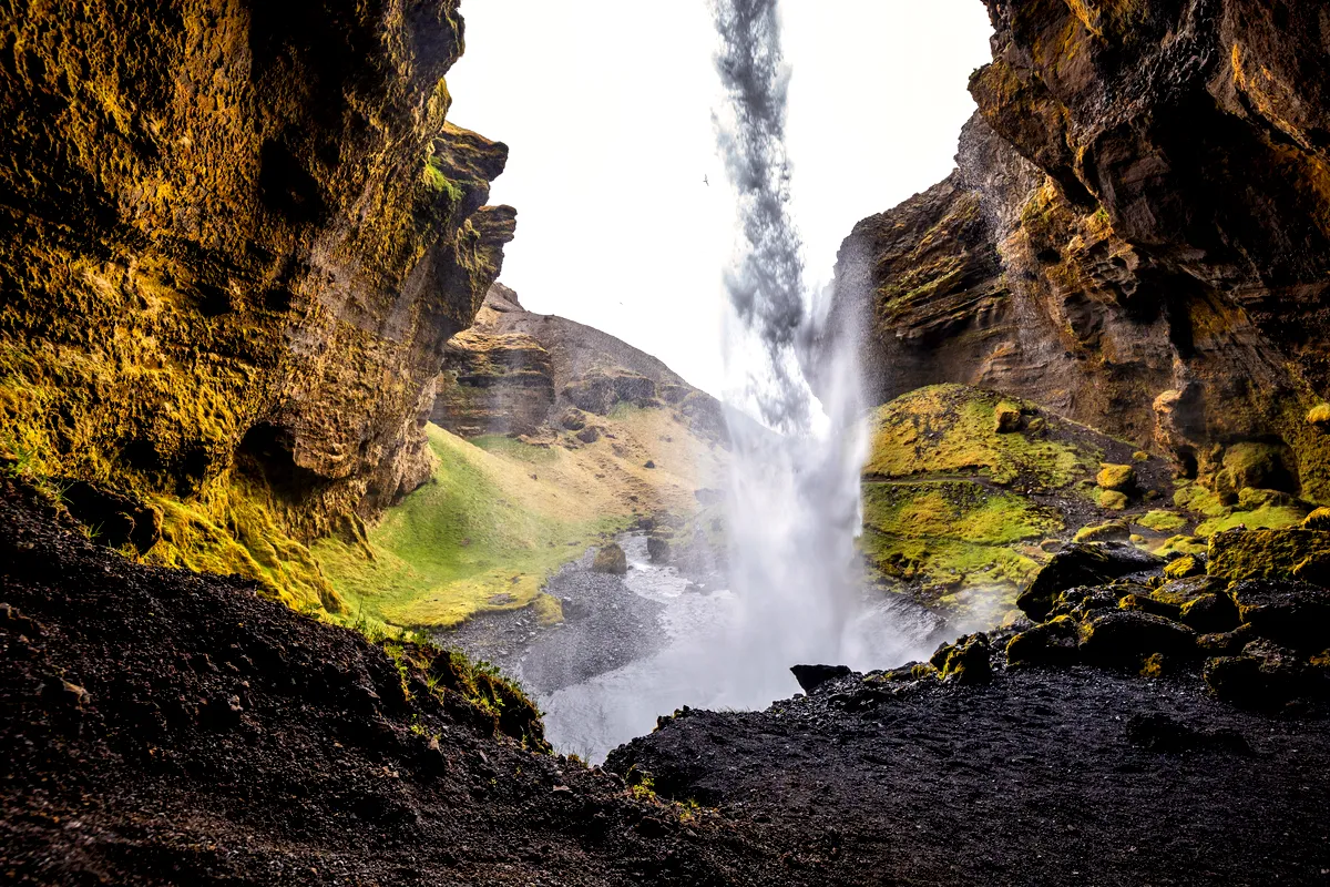 A breathtaking view of Kvernufoss Waterfall in Iceland, captured from behind the falls. The water cascades dramatically from a high cliff, creating a misty spray that fills the air. The scene is framed by rugged, moss-covered rock formations on either side, with lush green grass and patches of vegetation in the background. The ground is a mix of dark volcanic rocks and soil, adding to the contrast and beauty of the landscape. This image showcases the unique perspective and natural splendor of one of Iceland's hidden gems.