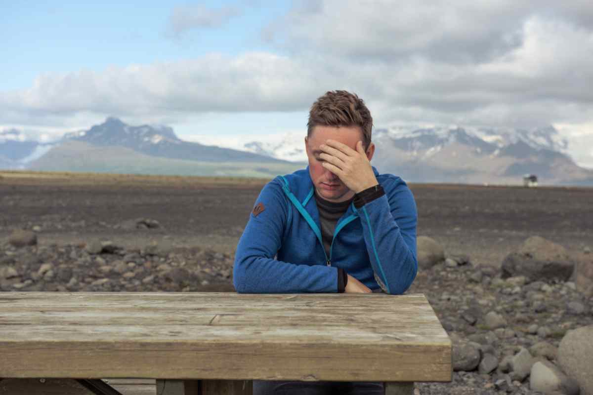 Tired traveler in a blue jacket sitting at a picnic table, head in hand, in a stark volcanic landscape.