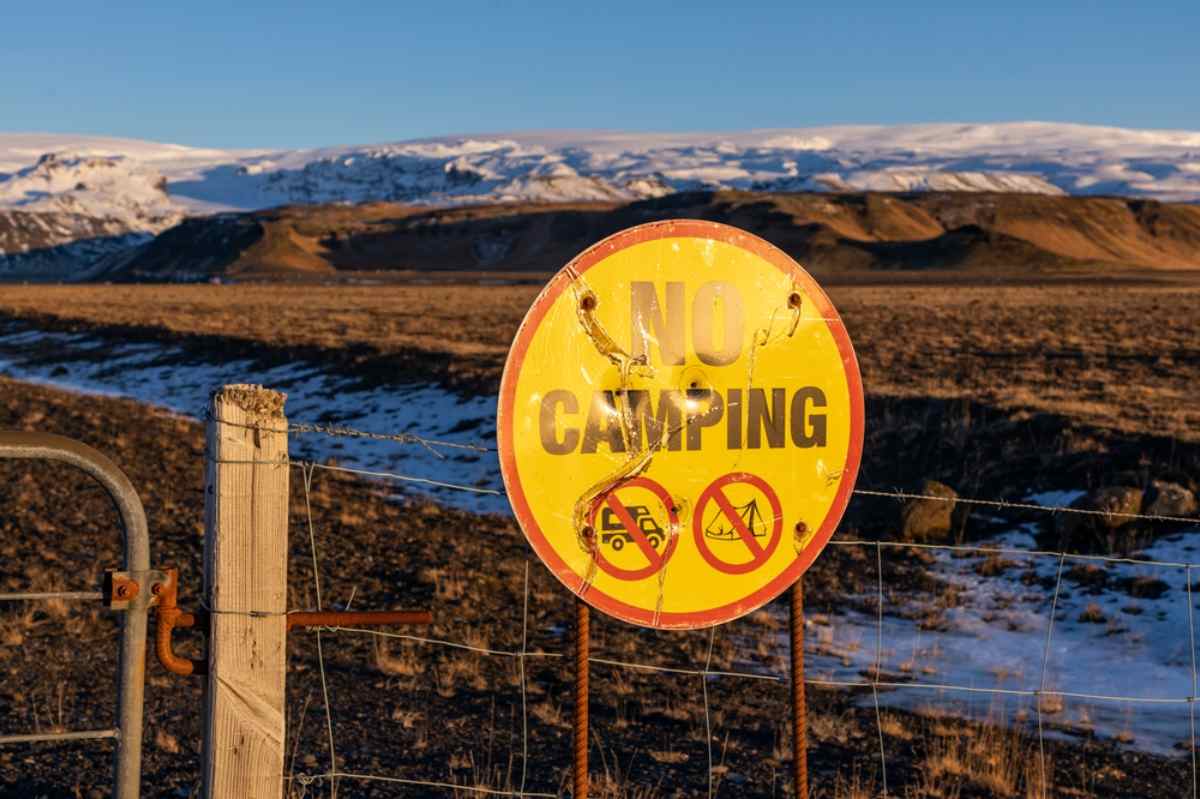 Weathered yellow “No camping” sign on a fence with snowy Icelandic mountains in the background.