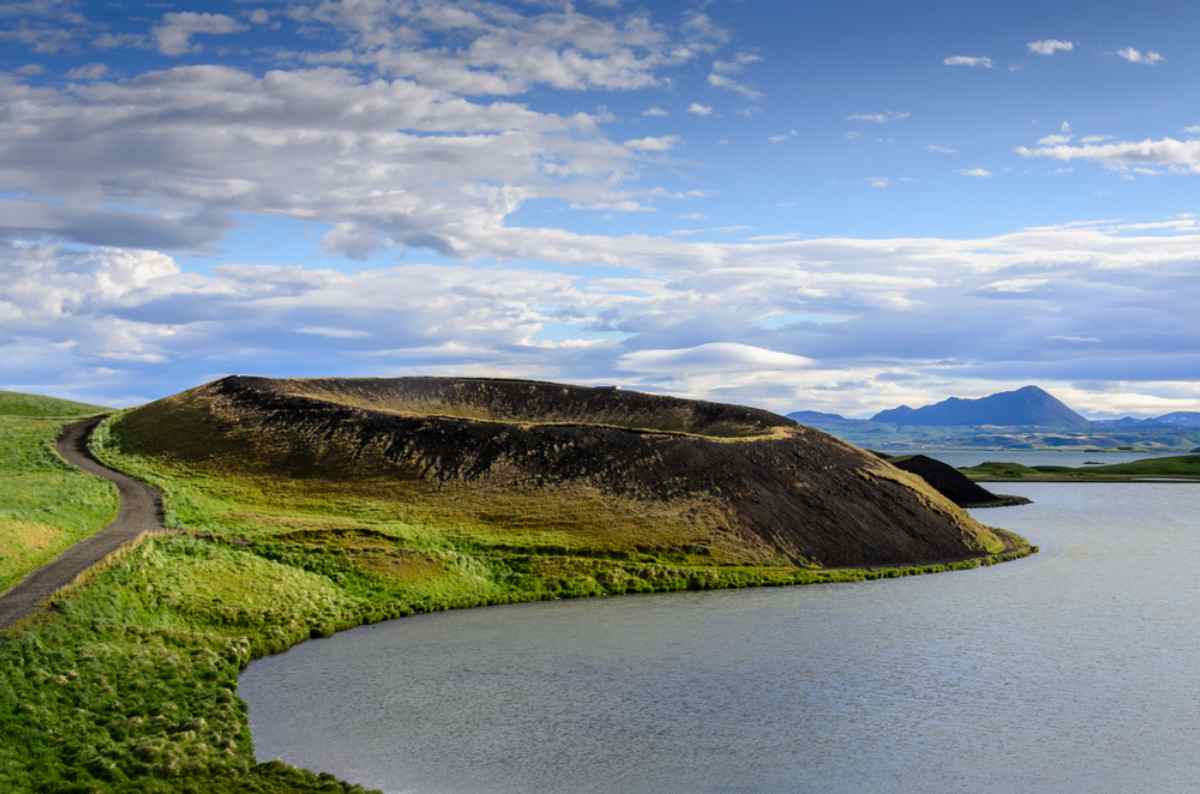 Walking path around a green pseudo-crater on the shore of a calm lake in North Iceland.