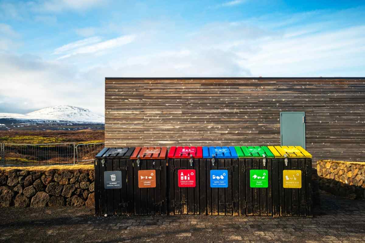 Row of color-coded recycling and trash bins in front of a wooden building with snowy hills behind.