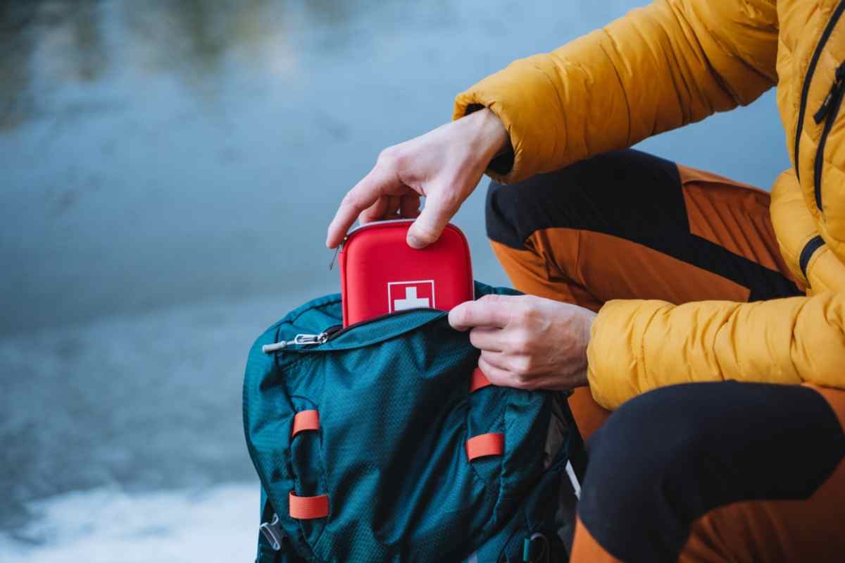 Hiker in a yellow jacket placing a red first-aid kit into the front pocket of a teal backpack by the water.
