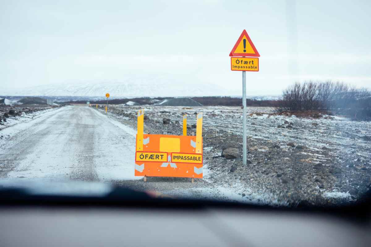 Consultar el tiempo y el estado de las carreteras en Islandia Señal de advertencia luminosa “Ófært / Impassable” y barrera en una carretera de grava nevada en Islandia, baja visibilidad y paisaje invernal por delante.