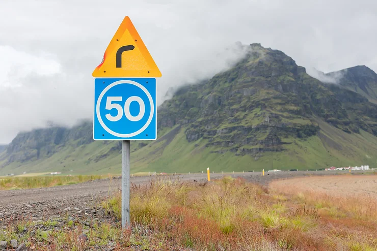 La importancia de conocer los límites de velocidad en Islandia Señal de tráfico en Islandia que muestra 50 km/h y una advertencia de giro a la derecha, con montañas cubiertas de musgo y nubes bajas de fondo en un día brumoso.