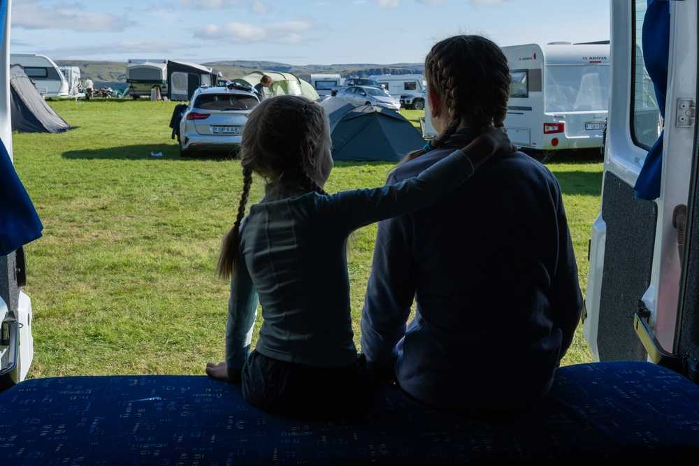 Two travelers sitting in a campervan doorway, looking out over a busy Iceland campsite with tents and RVs.