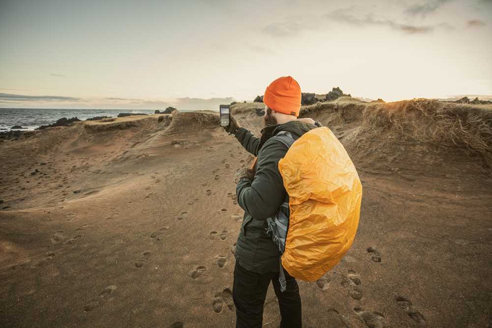 Traveler in an orange beanie checking their phone signal on a windy Iceland beach at sunset.
