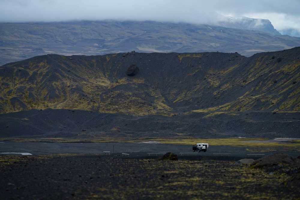 Camper driving through Iceland&rsquo;s black volcanic highlands with crater slopes and moody mountain weather.