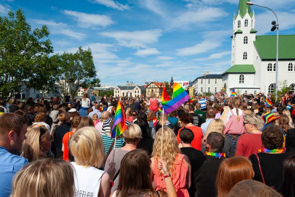 Crowds waving rainbow flags at Reykjavik Pride near Hallgr&iacute;mskirkja on a sunny day.