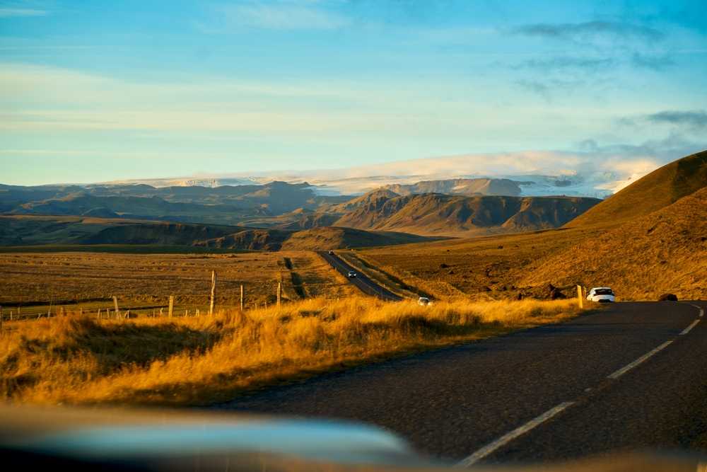 Scenic Ring Road drive through golden Icelandic hills with distant mountains under a clear summer sky.