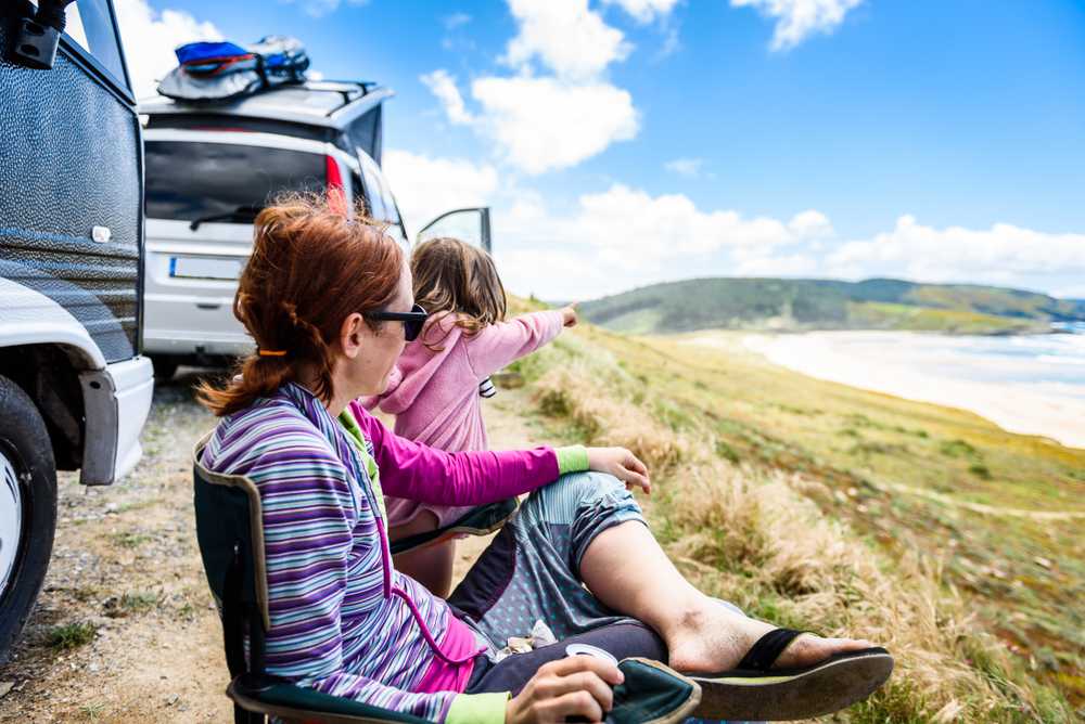 Traveler and child relaxing beside a campervan, looking out over an Icelandic coastline on a sunny day.
