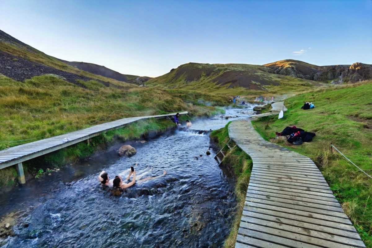 People bathing in a natural hot spring in Iceland beside wooden boardwalks and green hills