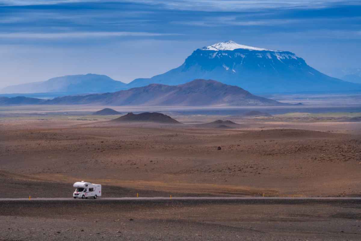 A white motorhome drives on an Icelandic road with a huge snow-capped mountain in the distance