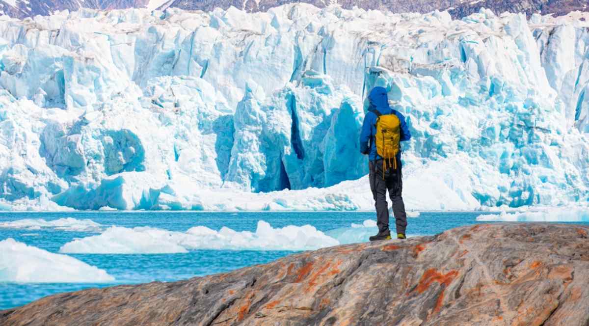 Solo hiker in bright gear standing on rocky ground facing a massive blue Icelandic glacier with floating icebergs on a calm summer day.