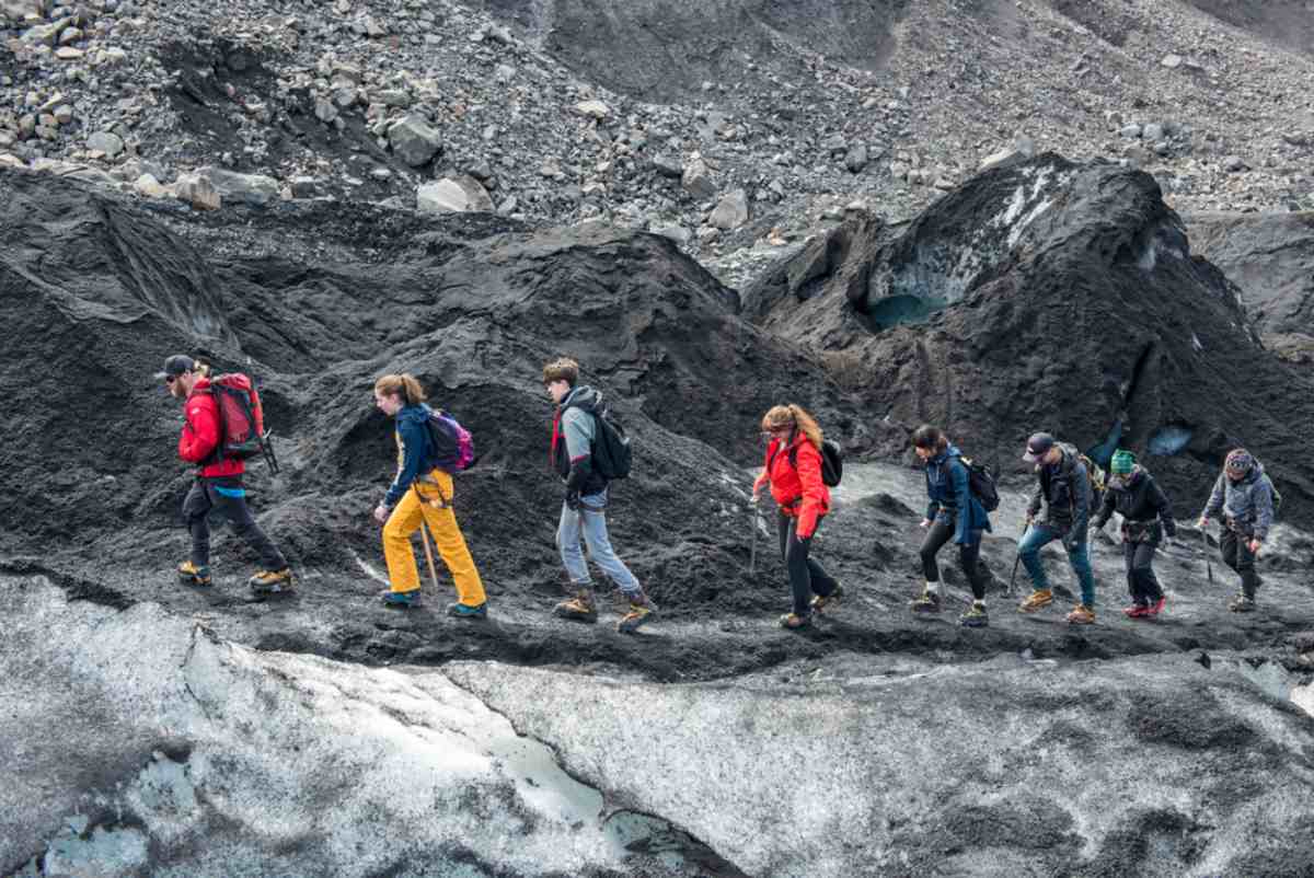 Group of hikers wearing helmets and winter gear trekking across a rugged, ash-covered Icelandic glacier during a guided adventure.