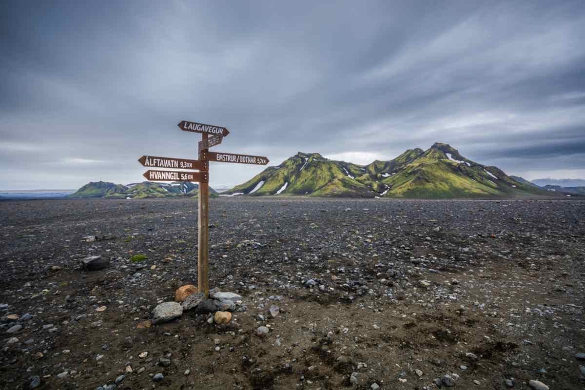 Wooden trail sign marking routes in Iceland’s Highlands, standing in a vast volcanic desert with green mountains under cloudy skies.