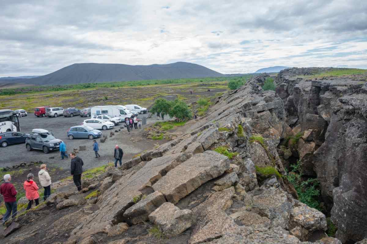 Visitors exploring an Icelandic lava rift near a busy parking area filled with cars and campervans on a cool summer day.