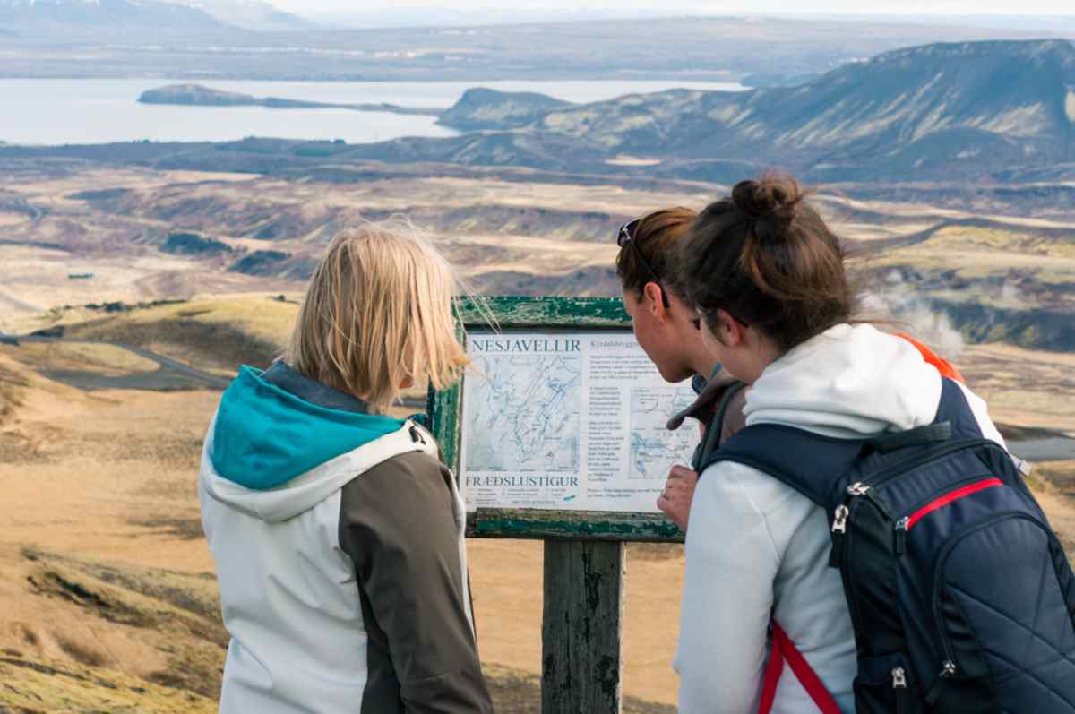 Tourists studying an outdoor map display overlooking wide Icelandic valleys, lakes, and mountains under a soft afternoon light.