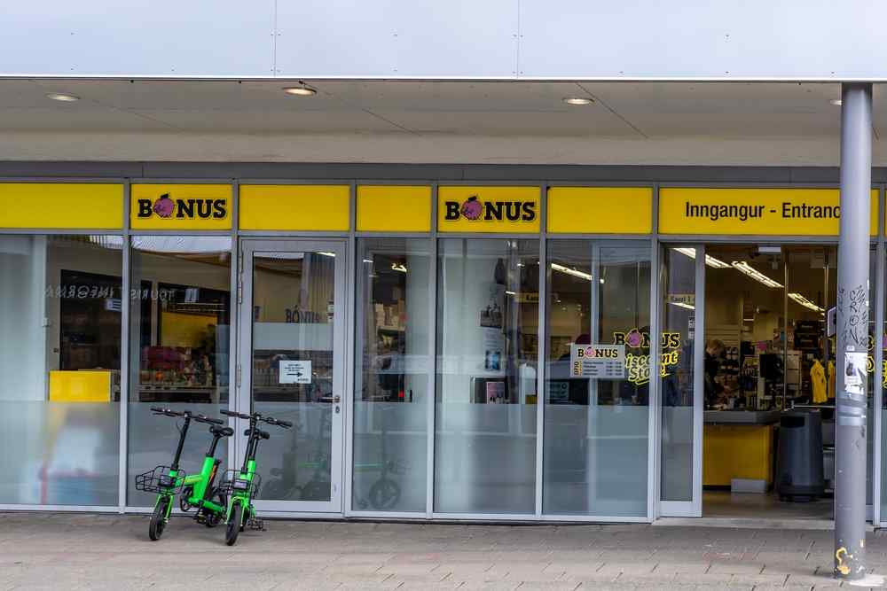 Entrance to a B&oacute;nus supermarket in Iceland with yellow signage and parked scooters outside.