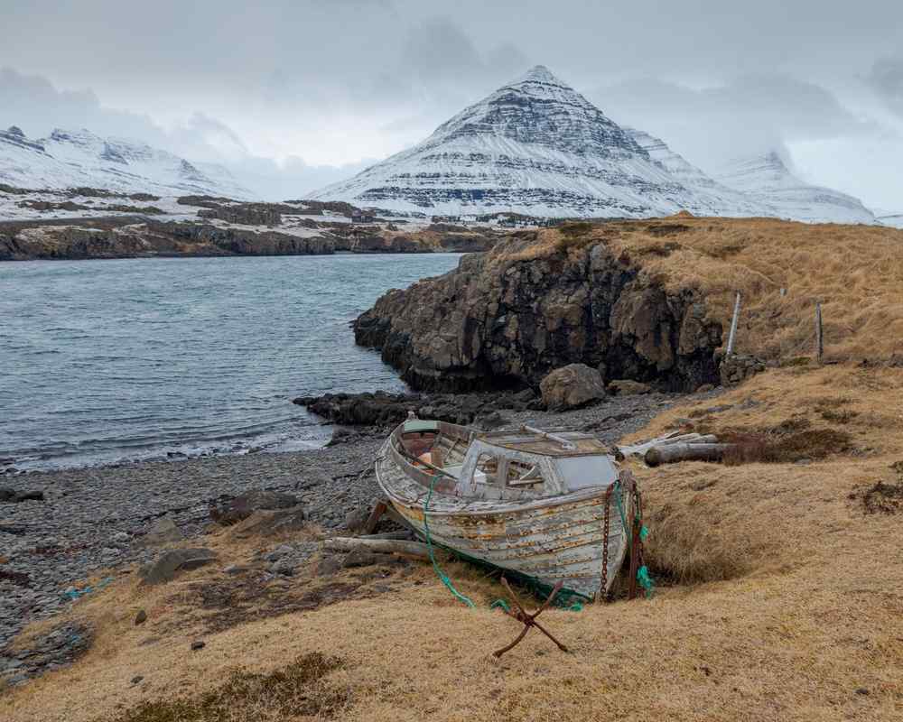 Old wooden boat on a rocky shore with a snow-capped mountain across the fjord.