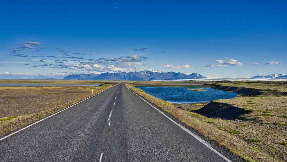 Empty Icelandic road stretching toward distant mountains under a clear blue sky.