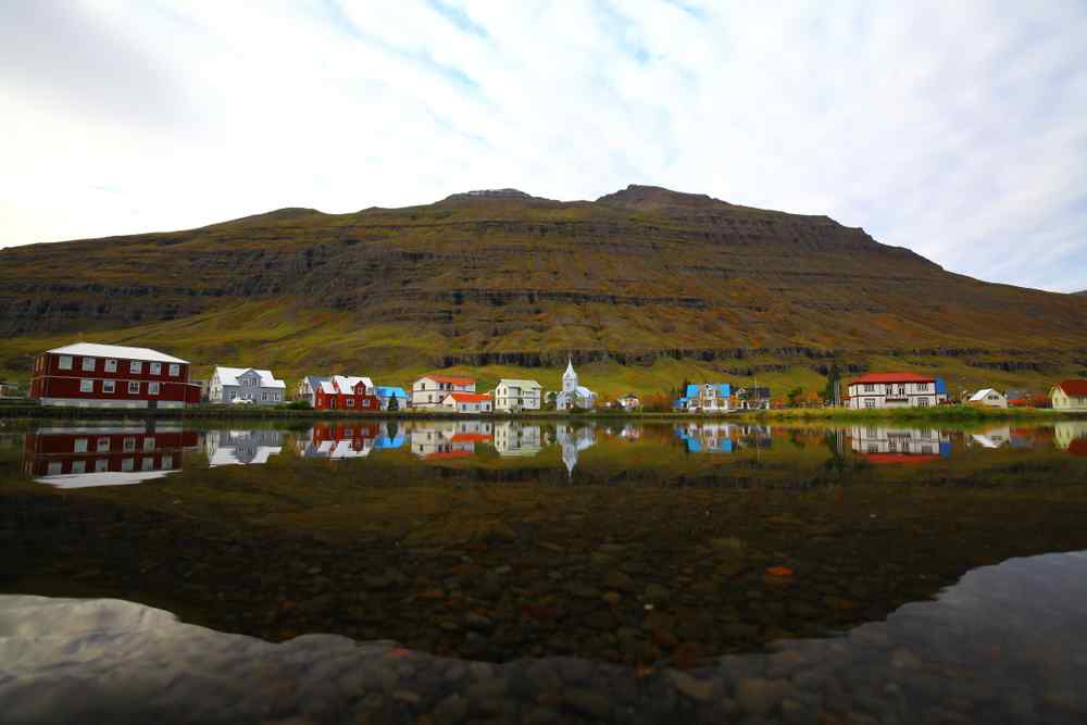 Colorful houses in Sey&eth;isfj&ouml;r&eth;ur reflected in calm water beneath a steep mountain.