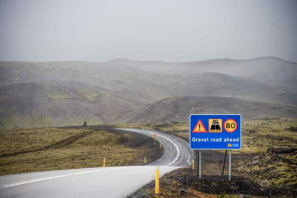 Gravel road ahead sign beside a rural Icelandic road in misty hills.