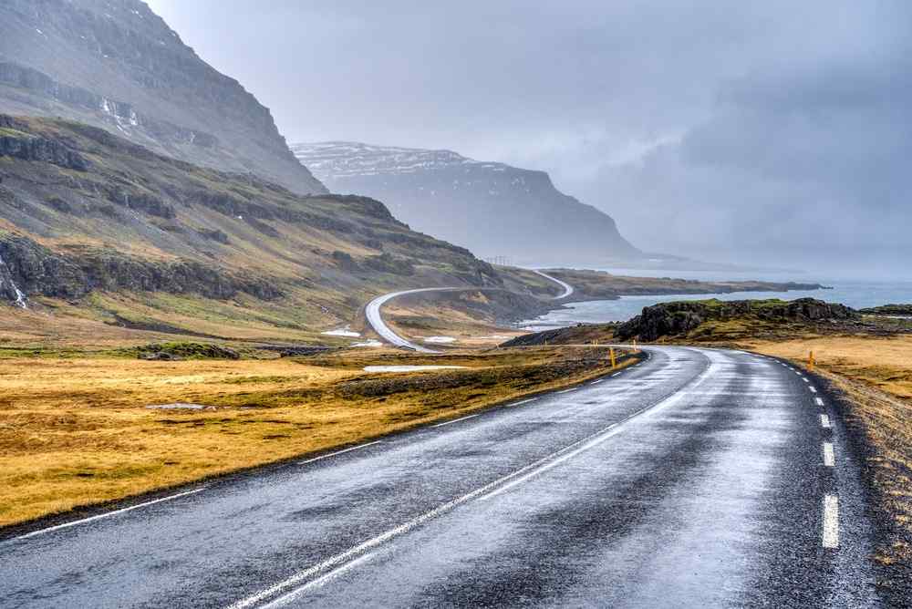 Wet Icelandic road winding through dramatic coastal mountains under cloudy skies.